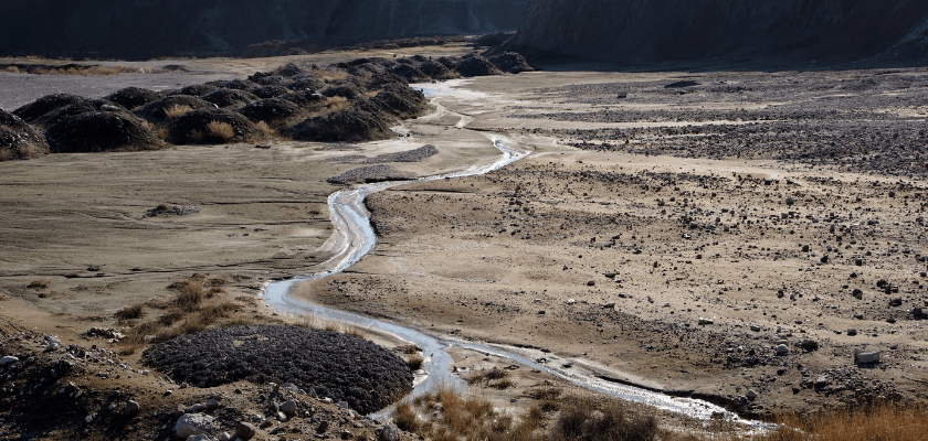 Small water channel cutting through arid, sandy terrain with dark mounds of earth and distant hills.