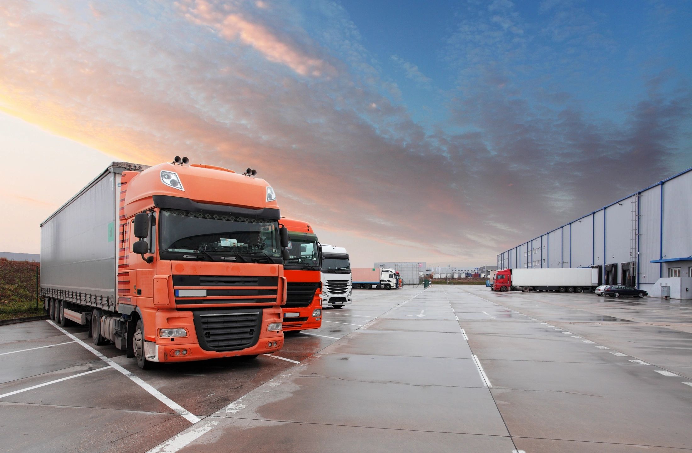Orange and white freight trucks parked in a logistics center loading dock during sunset, representing supply chain and transportation services.