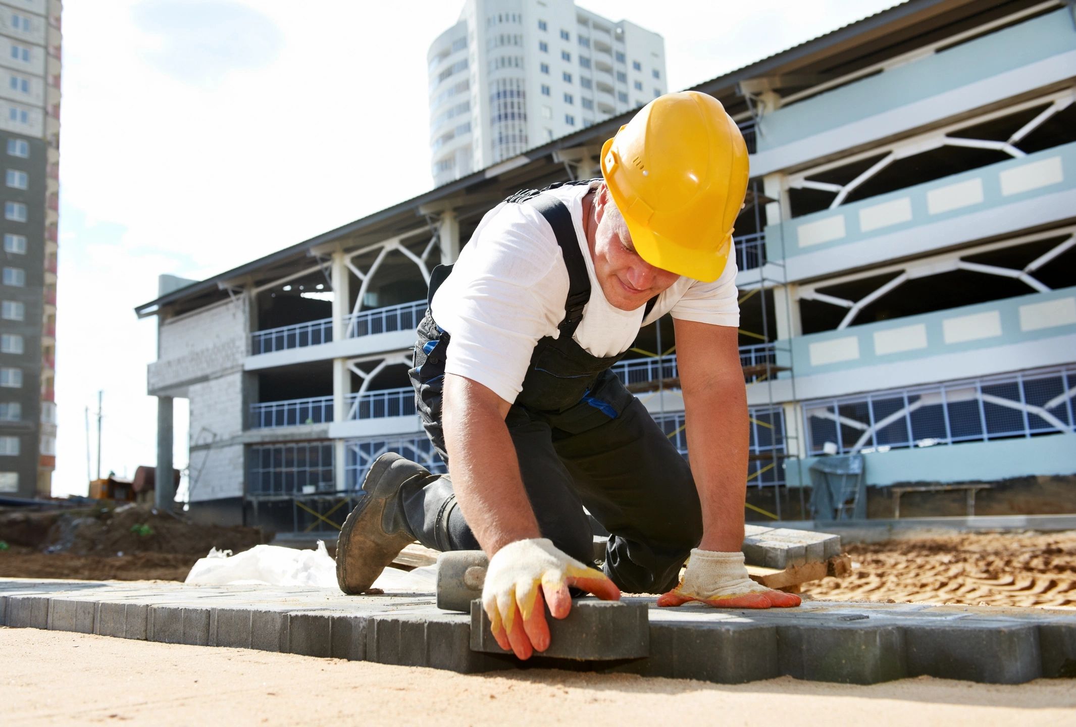 Construction worker installing gray interlocking patio pavers at a commercial construction site with modern buildings in the background.