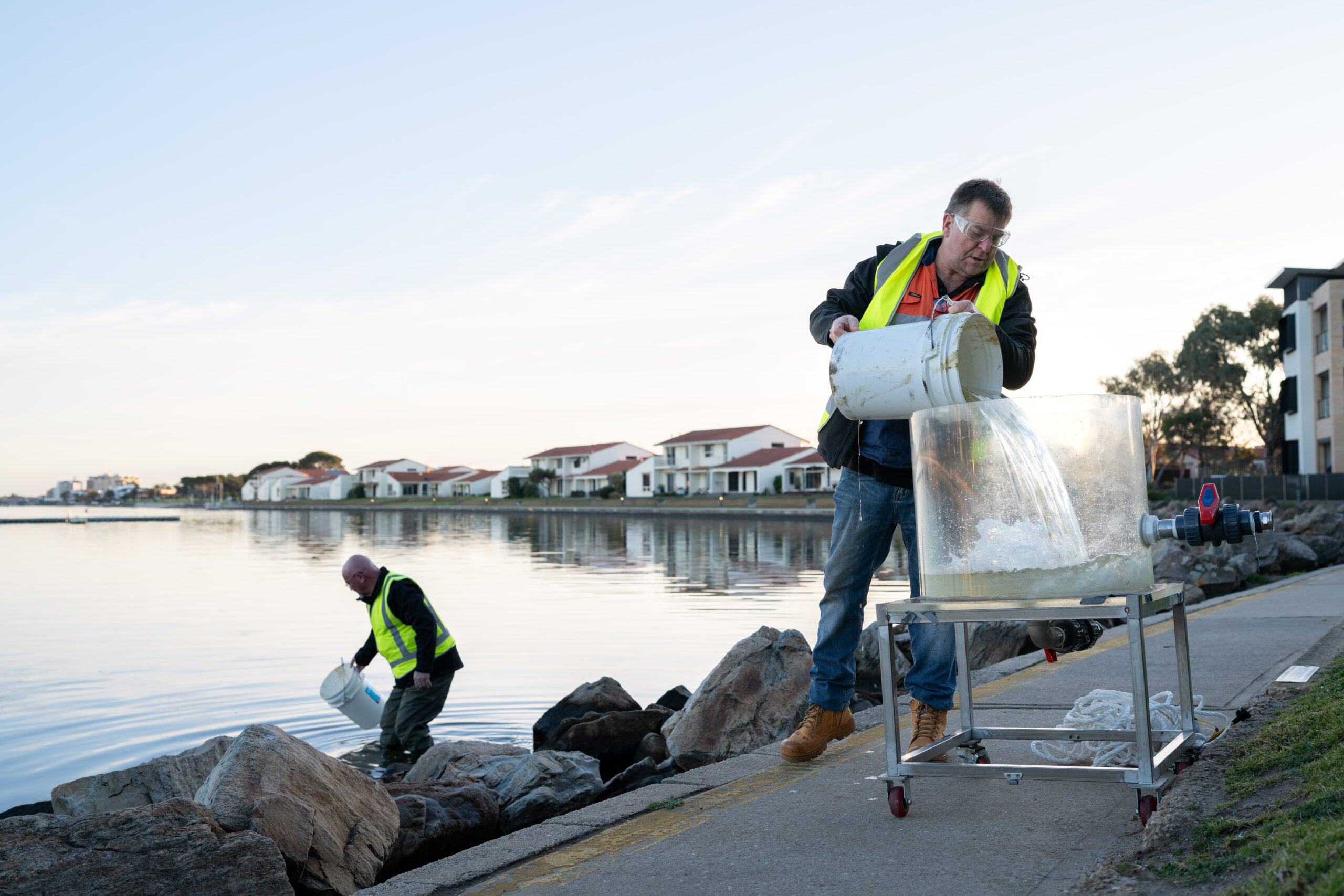 Environmental technicians collecting and processing water samples beside a lake, using buckets and filtration equipment for water quality testing.