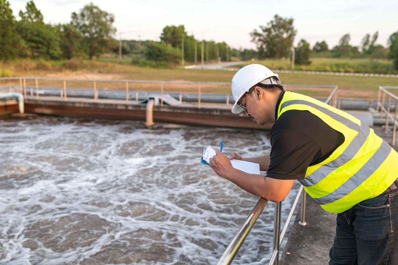 Water treatment plant worker wearing a hard hat and safety vest inspecting aeration tanks and recording measurements during wastewater treatment.